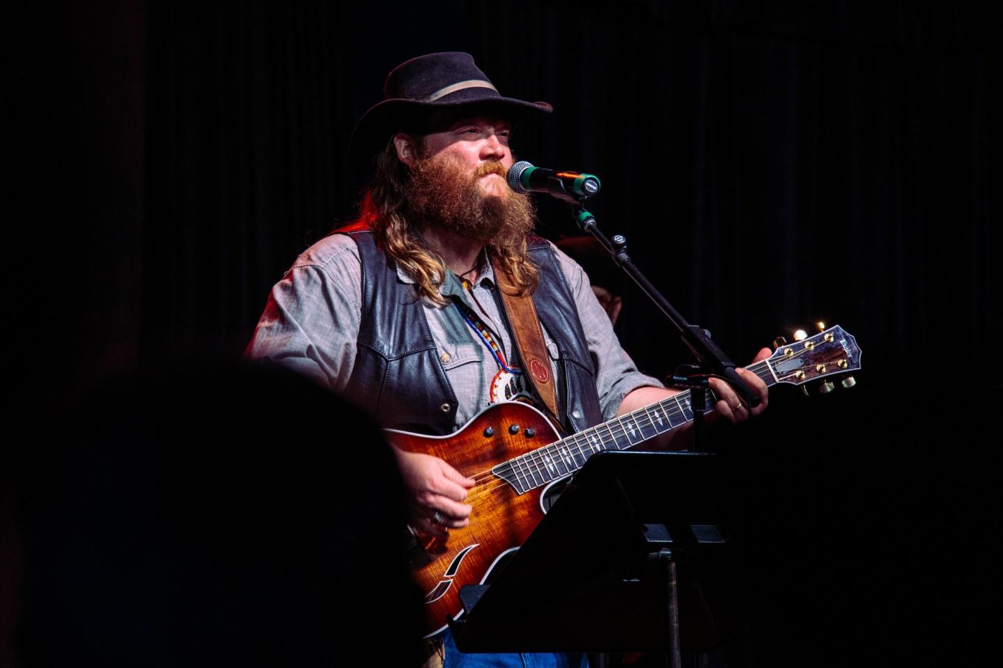 Musician with guitar performing on stage, wearing a hat and vest.