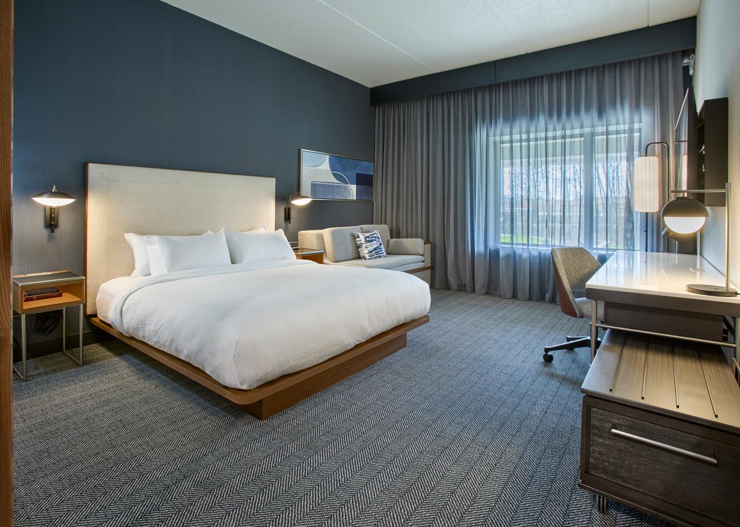 Modern hotel room with a king bed, seating area, desk, and large window with curtains.