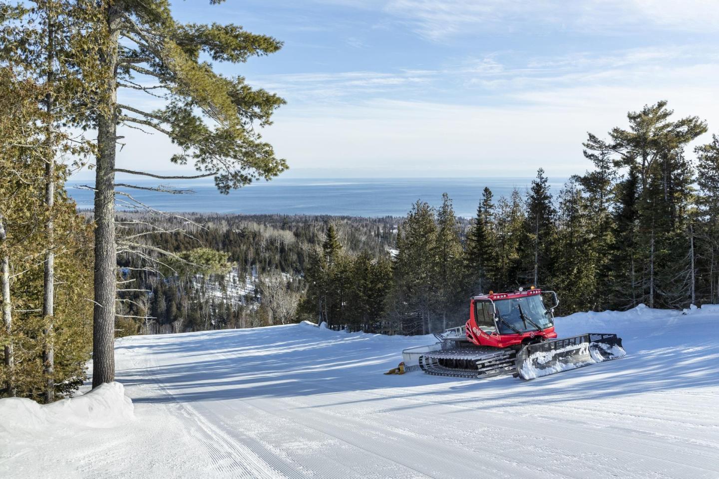 Snow groomer on a snowy slope with a forest and sea in the background.