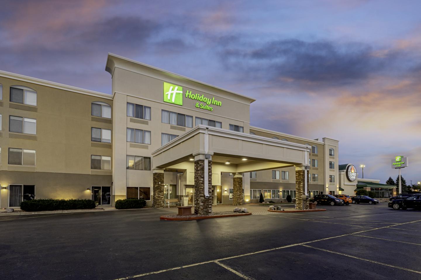 Modern hotel exterior at dusk with glowing signs and ample parking.
