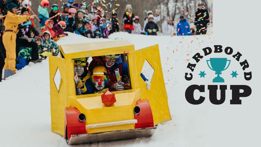 Cardboard car sled with riders in costumes, spectators watch on snowy hill.