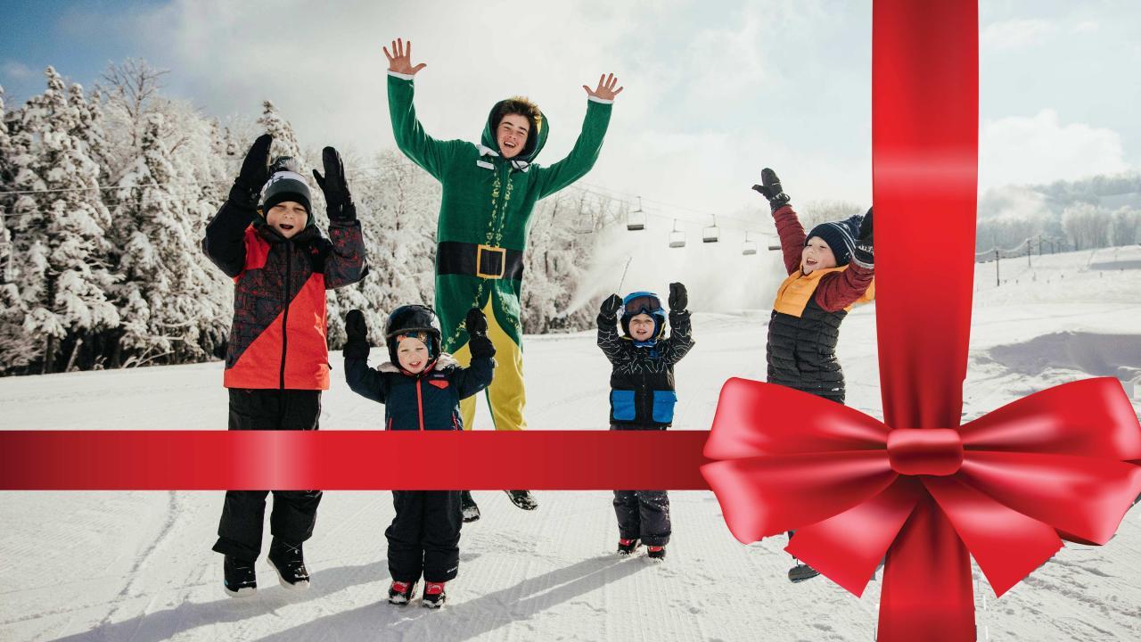 Snowy scene with five joyful people, one in a green outfit jumping. Large red ribbon decoration.