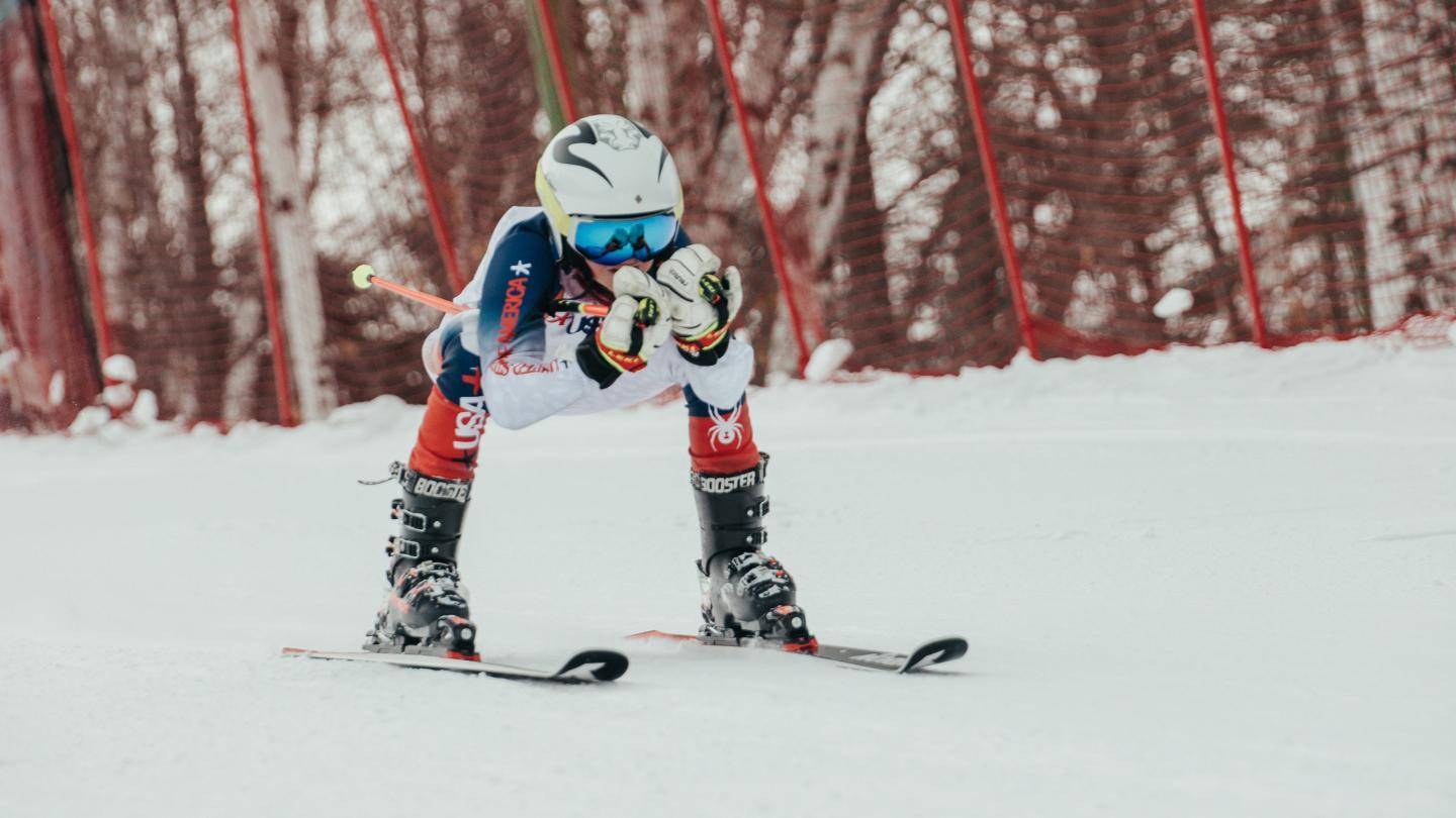 Young skier in racing pose on snowy slope, red fencing in background.