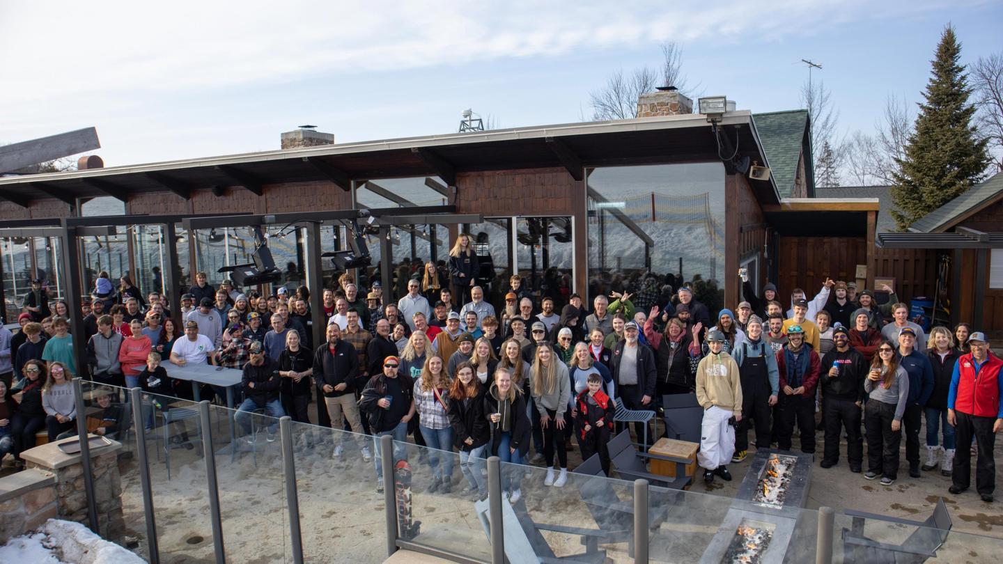 Large group of people gathered outdoors in front of a glass building on a clear day.