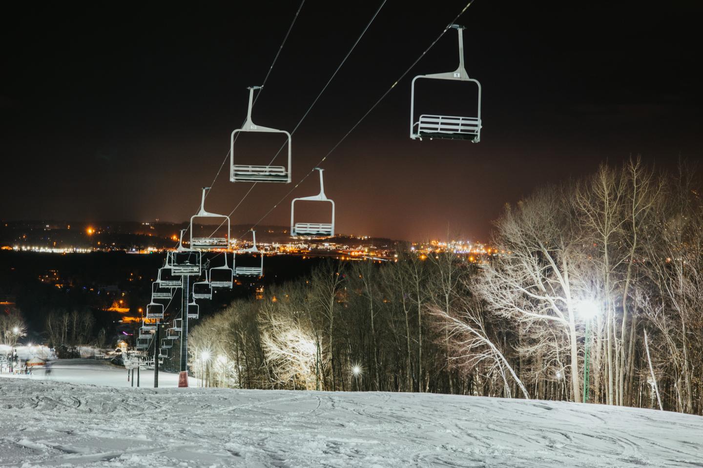 Ski lift chairs suspended, nighttime snowy landscape, city lights in distance.