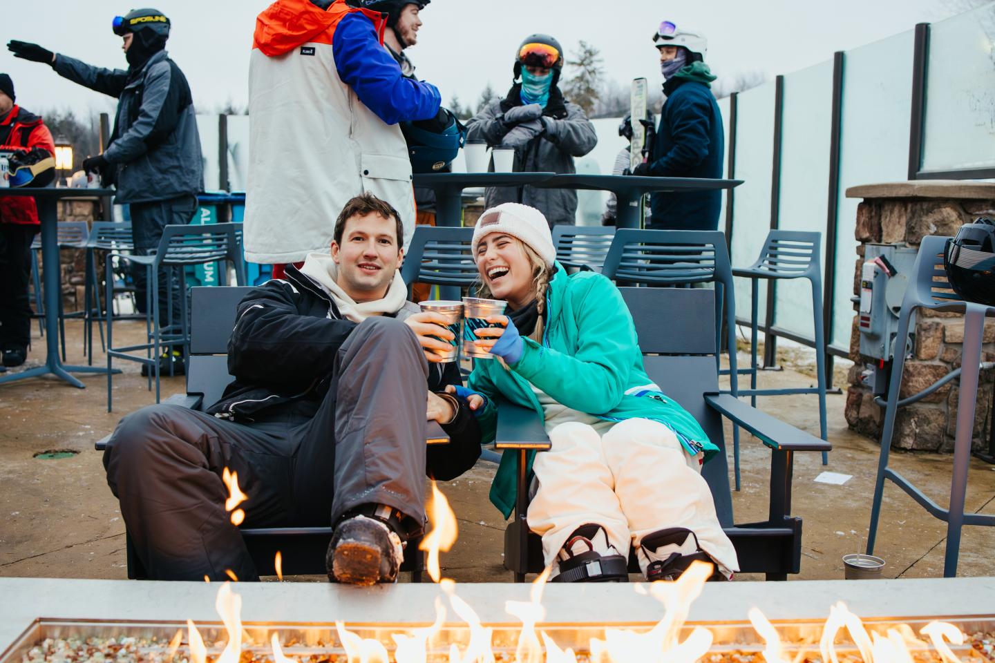 Couple in winter gear enjoys drinks by a fire, surrounded by other people in ski outfits.