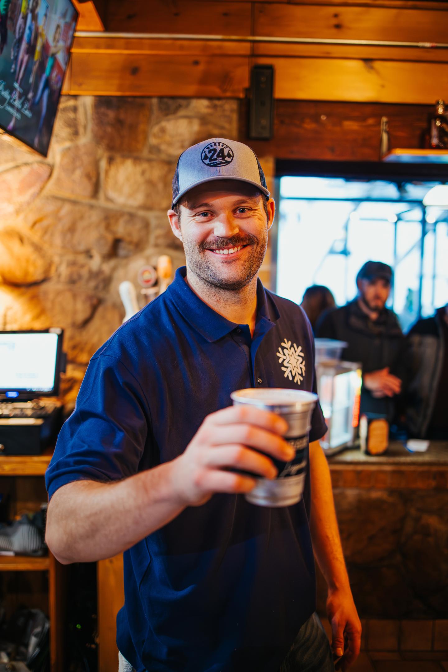 Smiling man in a cap offers a cup in a rustic wooden bar.