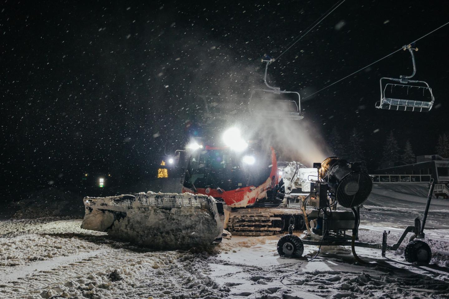 Snow grooming machine working at night on a snowy slope with ski lift nearby.