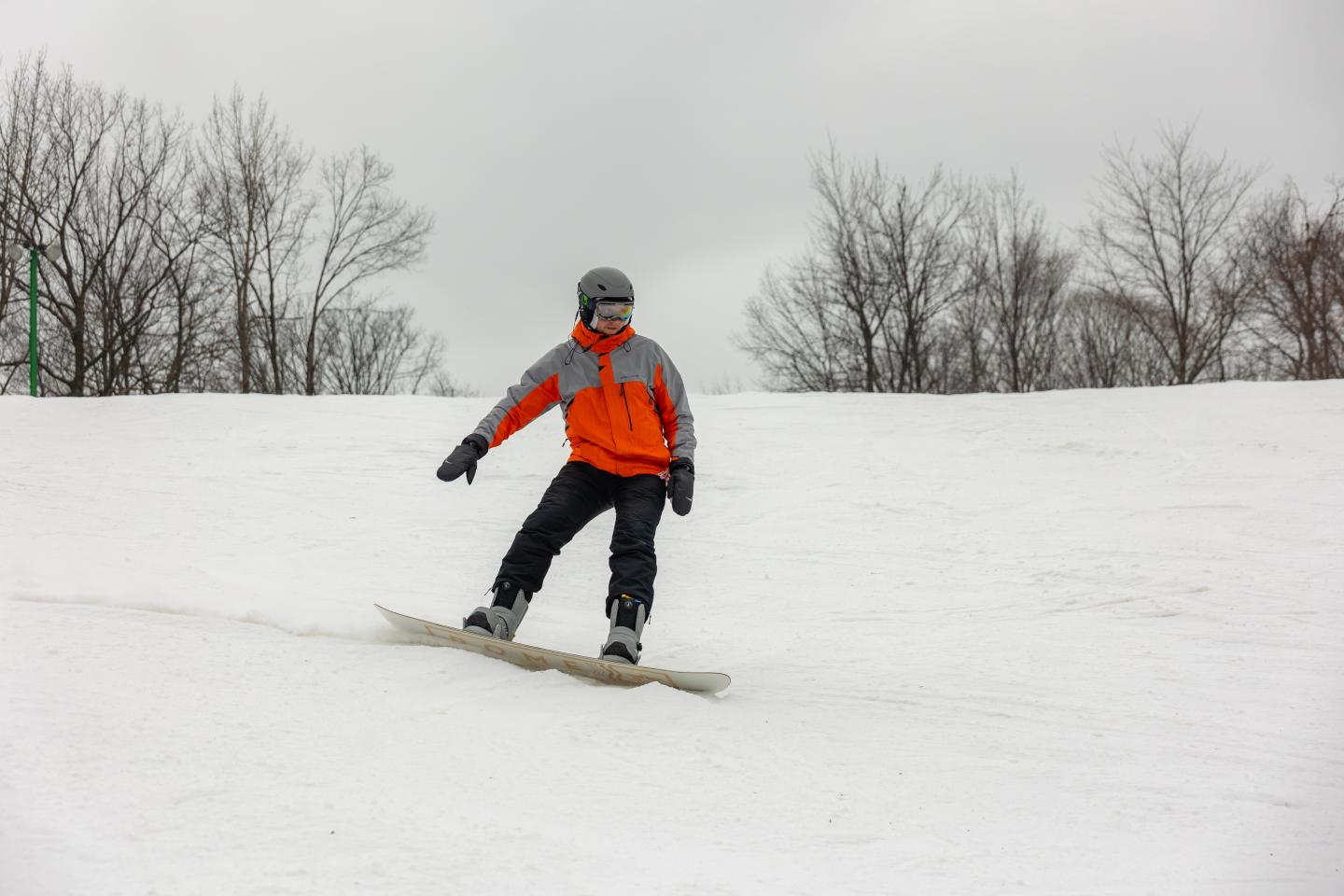 Snowboarder in an orange jacket glides down a snowy slope on a cloudy day.