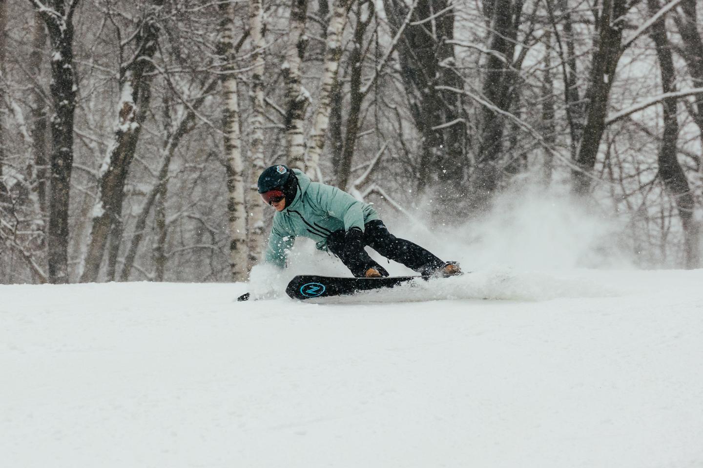 Snowboarder carving down a snowy slope amid winter trees.