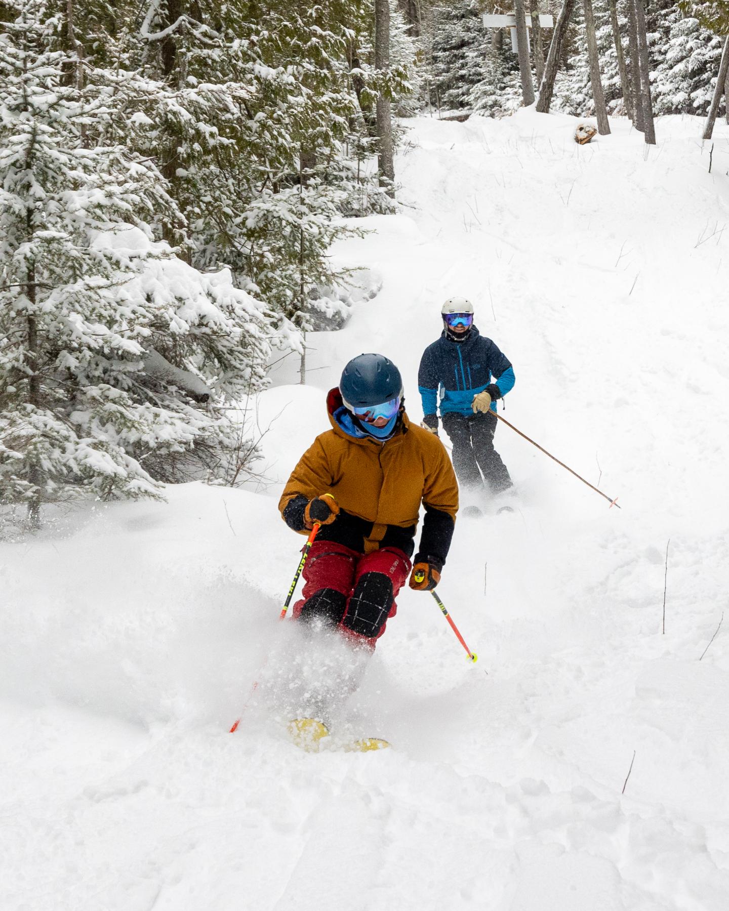 Two skiers wearing helmets and goggles on a snowy forest trail.