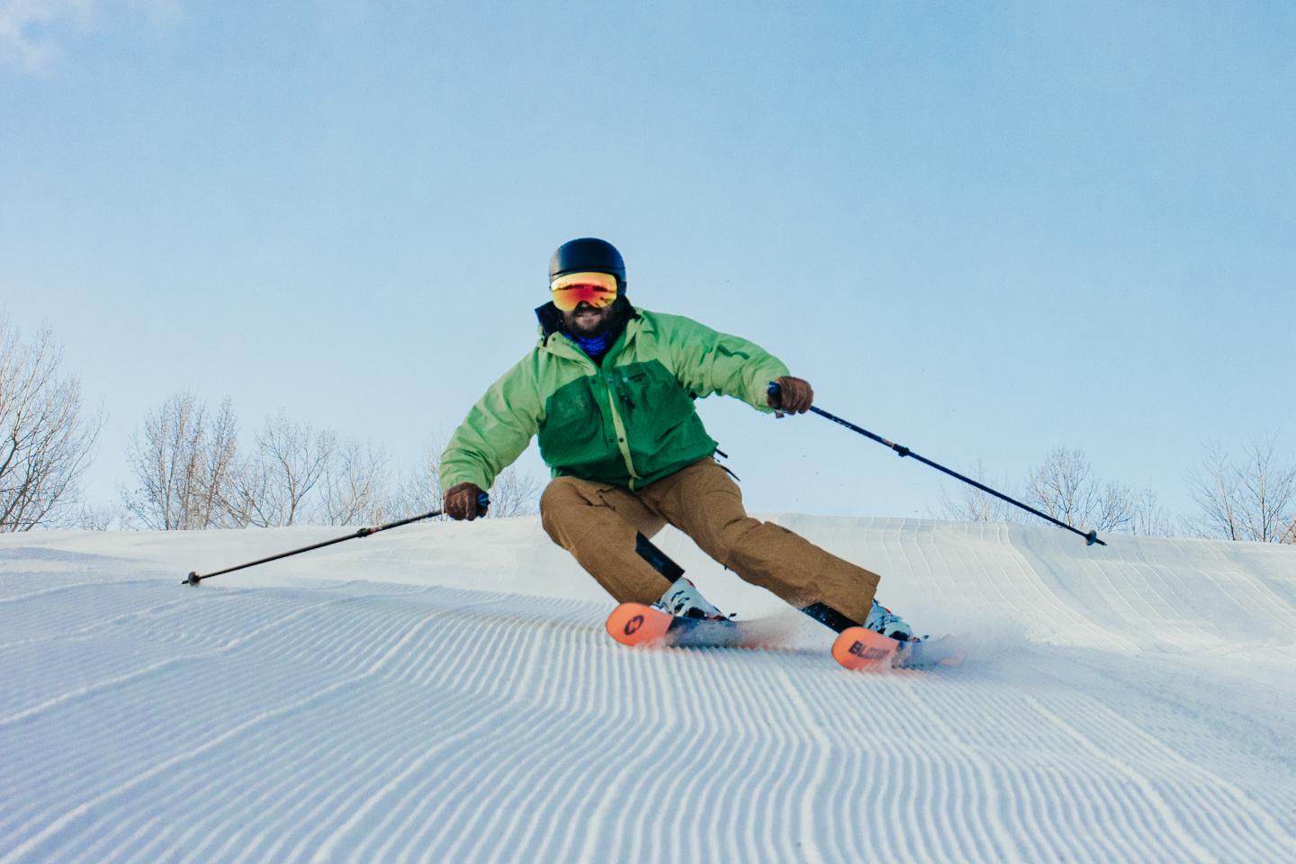 Skier in green jacket carving down a snowy slope under clear blue sky.