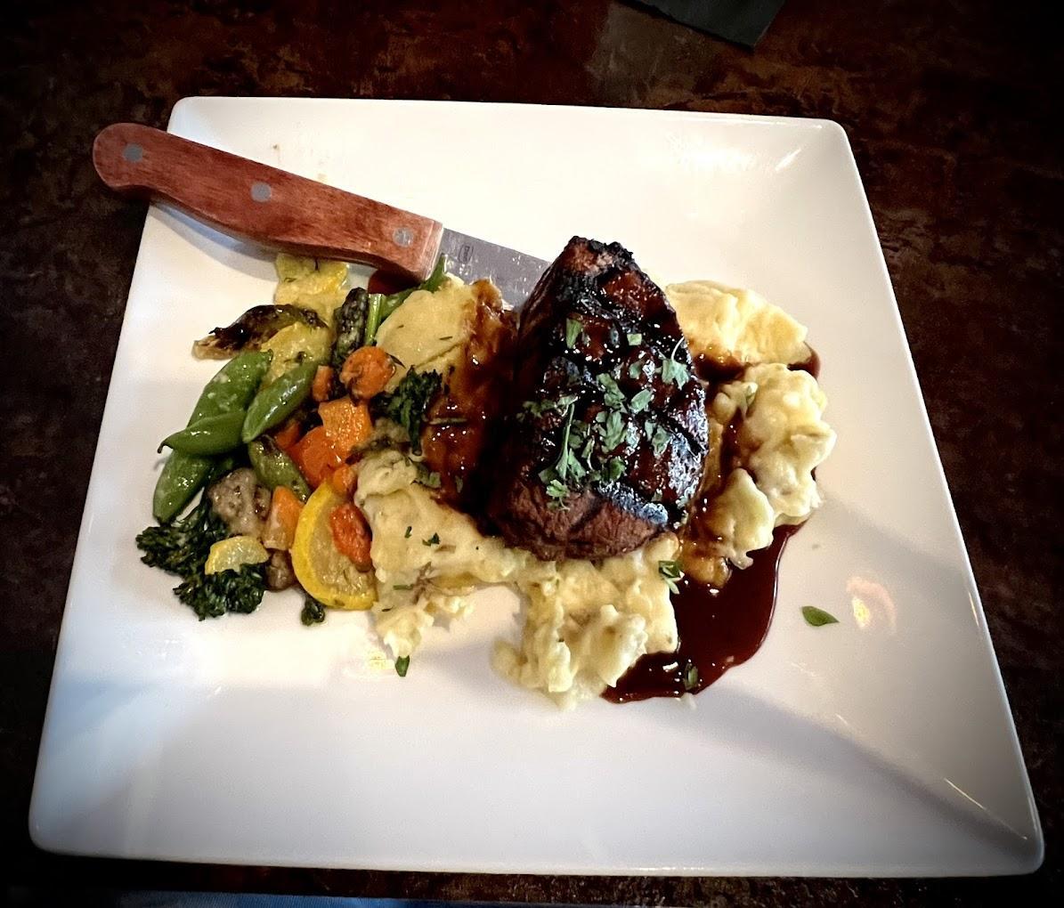 Steak with vegetables and mashed potatoes on a square plate, garnished with herbs.