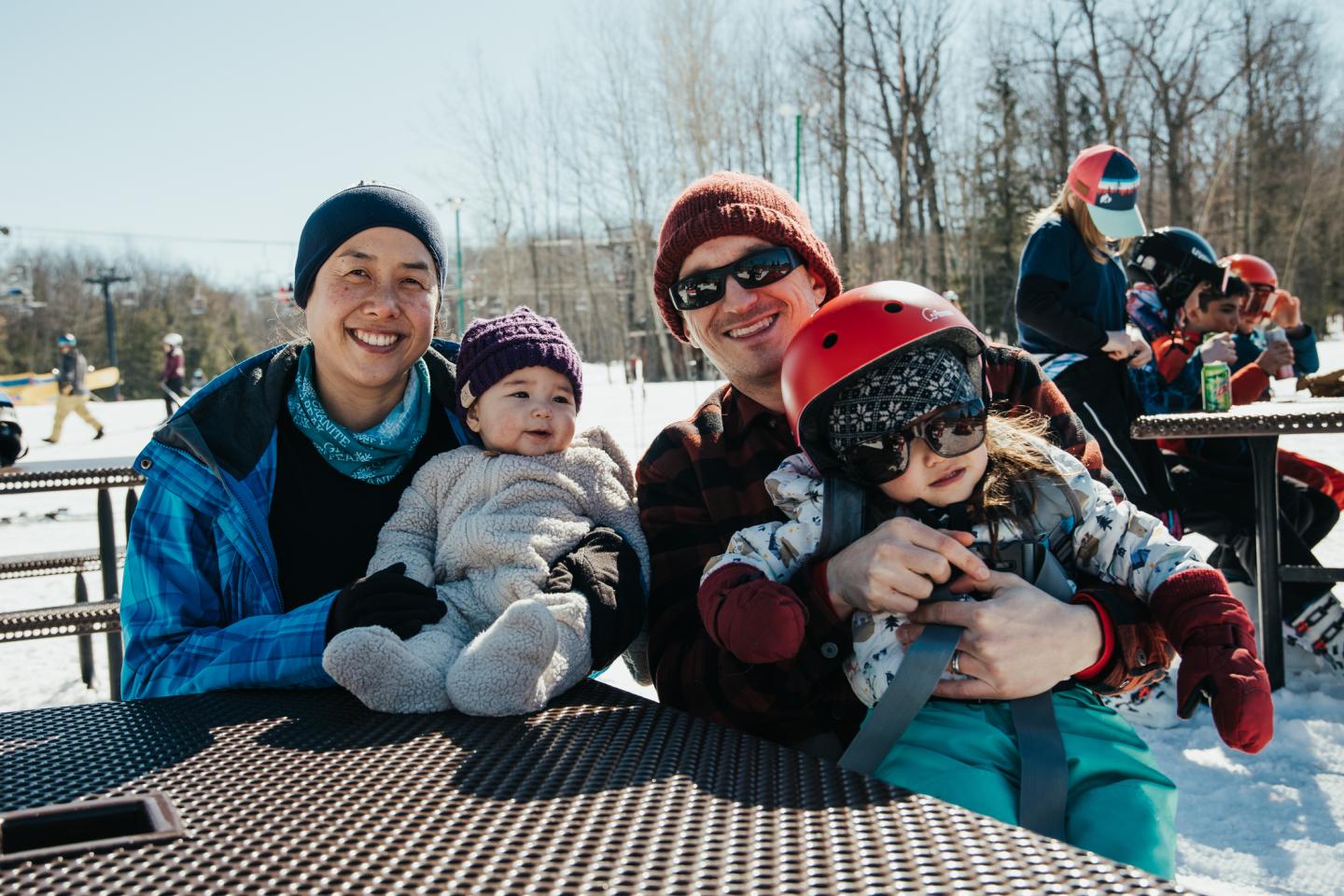Family sitting at an outdoor table in snow, smiling; kids wearing winter gear.