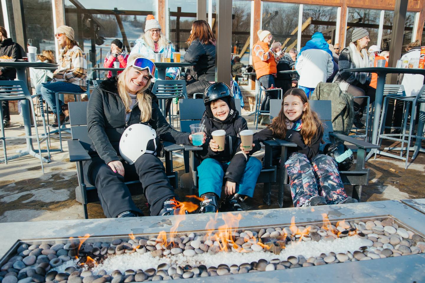 Three people in ski gear enjoying drinks by an outdoor fire pit.
