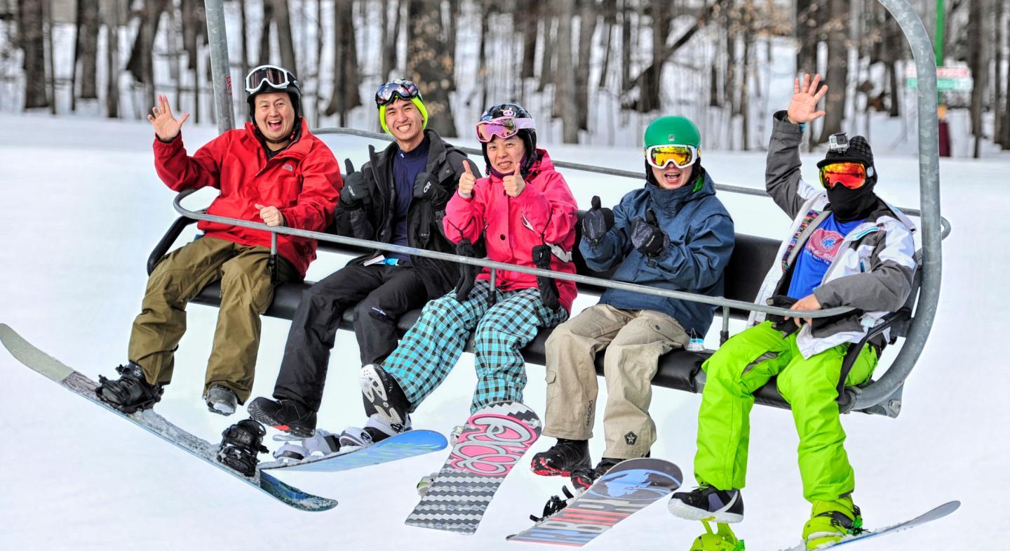 Five people on a ski lift, smiling and waving, with snowboards and snowy trees in the background.