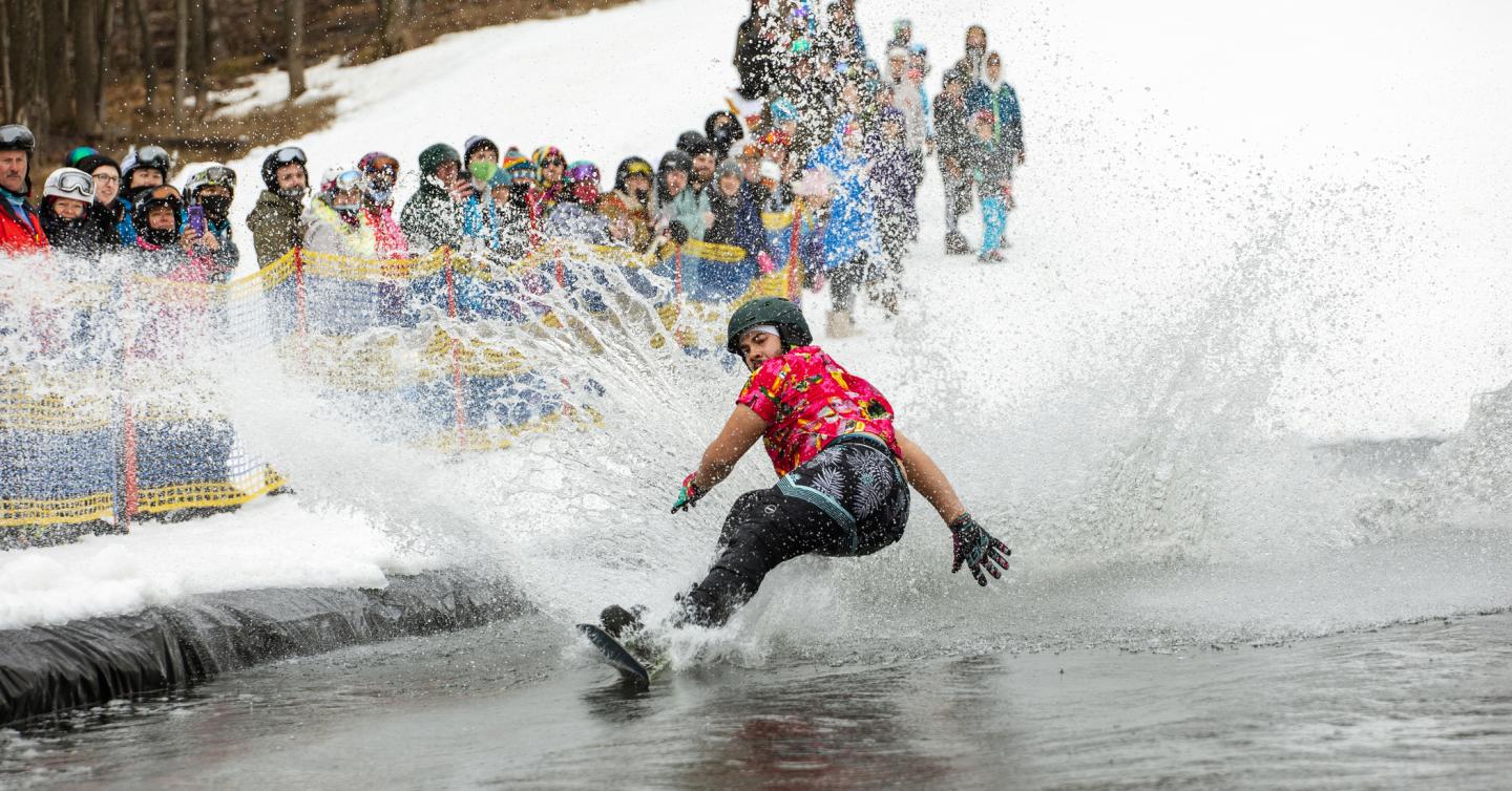 Snowboarder splashing through water with spectators watching.