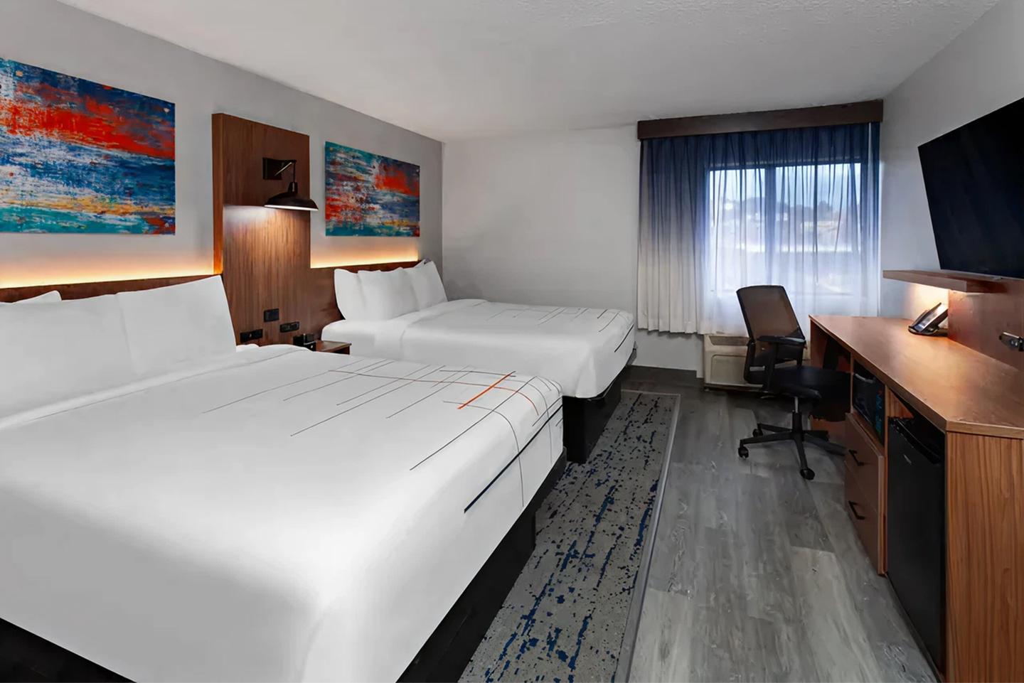 Two hotel beds, desk with chair, large TV, and abstract art on walls.