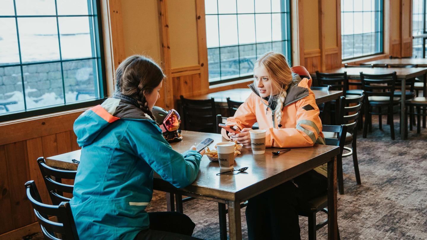 Two people in winter jackets chat at a table by large windows.