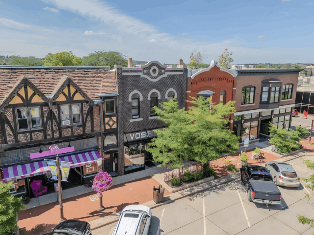 Street view of colorful shops with parked cars and trees under a blue sky.