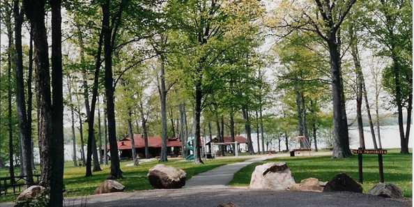 Pathway through a park with trees, large rocks, and a red-roofed pavilion near a lake.