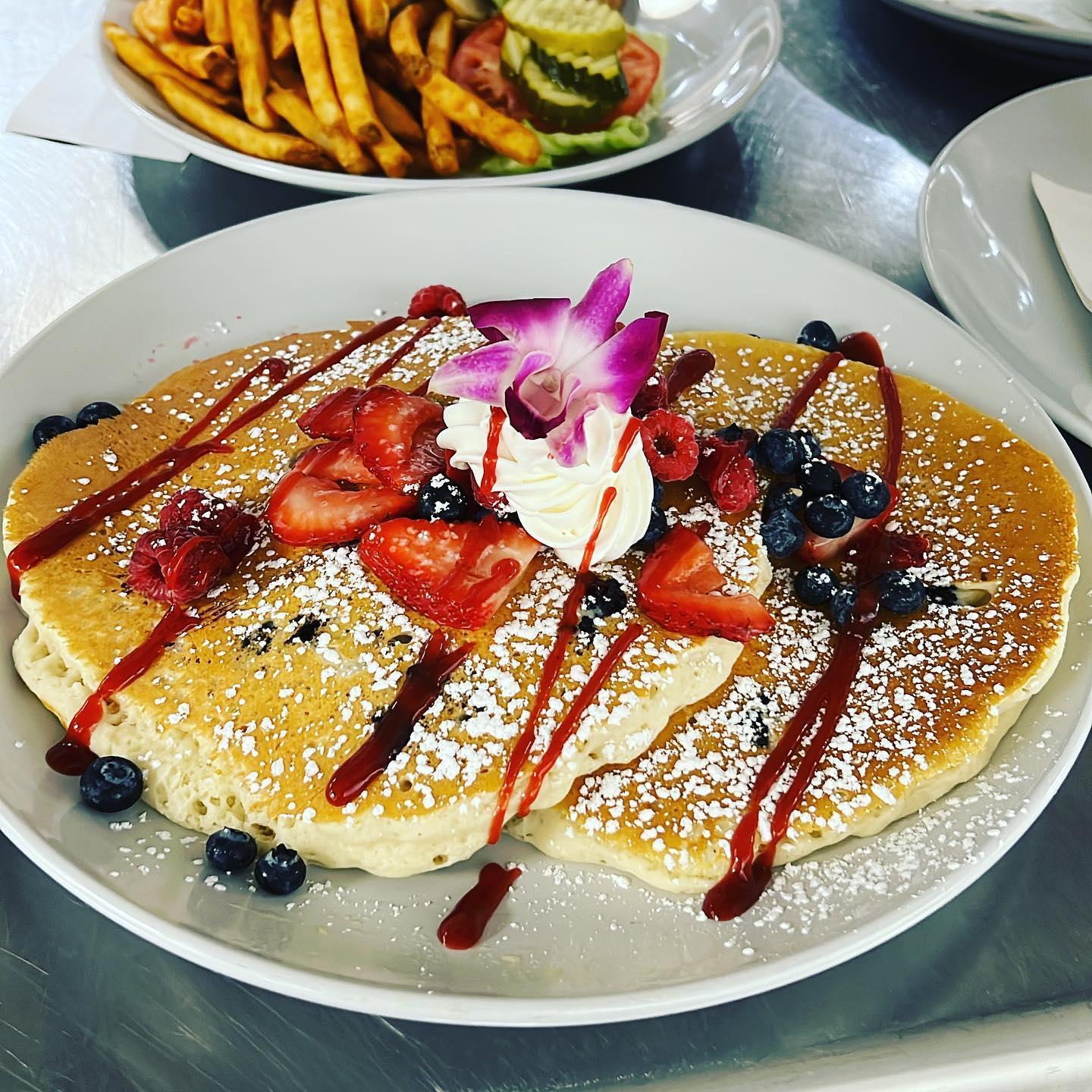 Pancakes with berries, whipped cream, and an orchid garnish. Fries and pickles in the background.