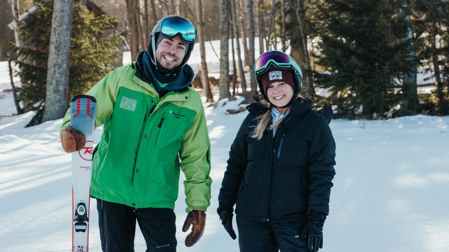 Two skiers smiling in winter gear, standing on snowy ground with trees in the background.