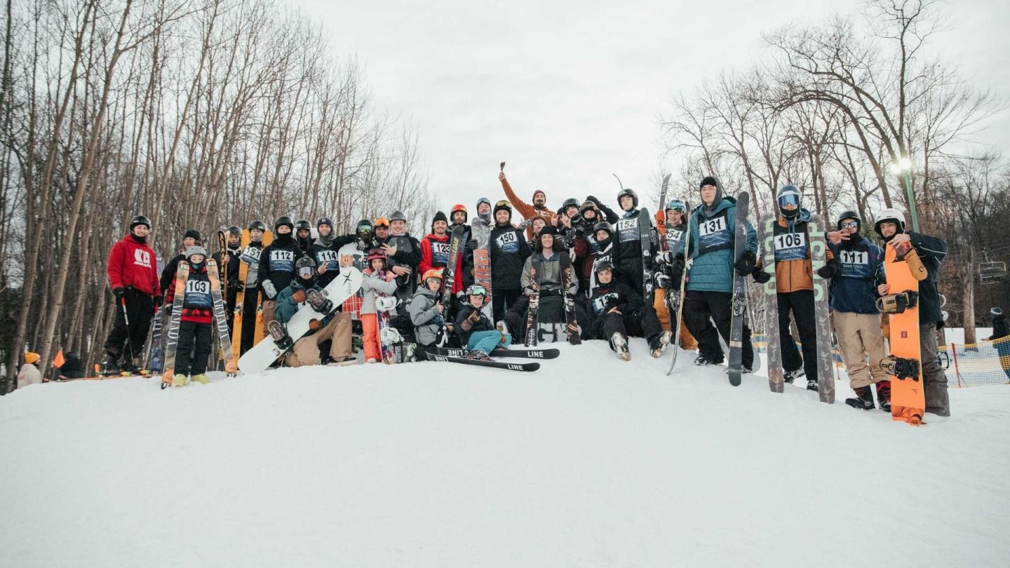 Snowboarders gathered on a snowy hill, surrounded by bare trees.