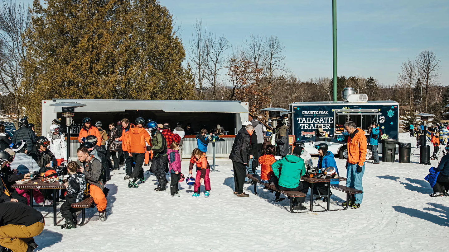 People in winter clothing gather at snow-covered outdoor seating near food trucks.
