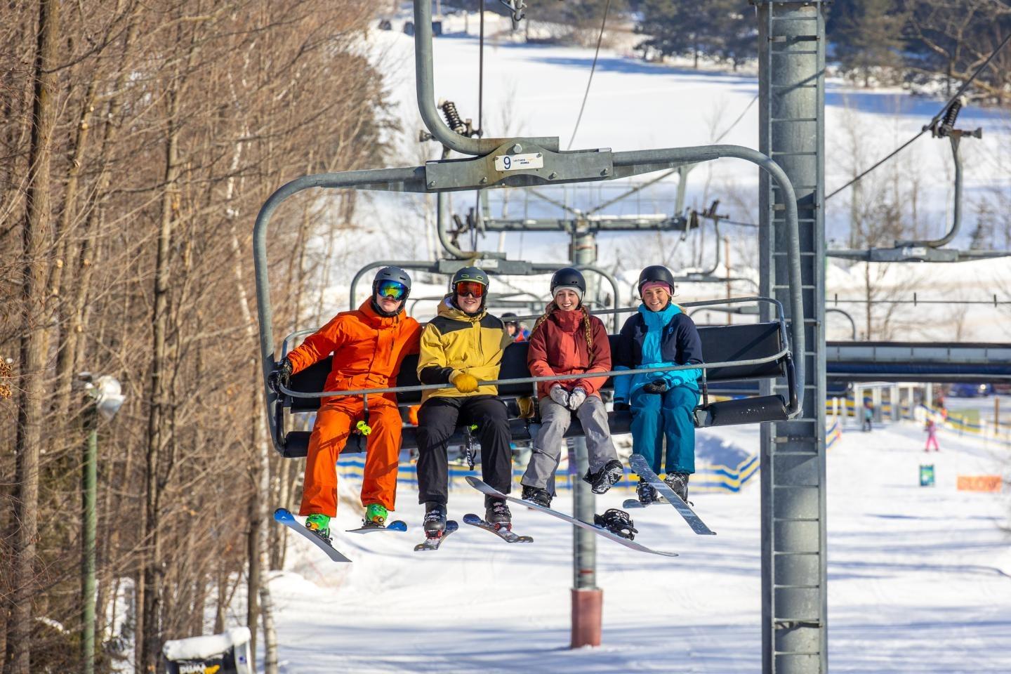 Skiers in colorful outfits on a chairlift with snowy trees behind.