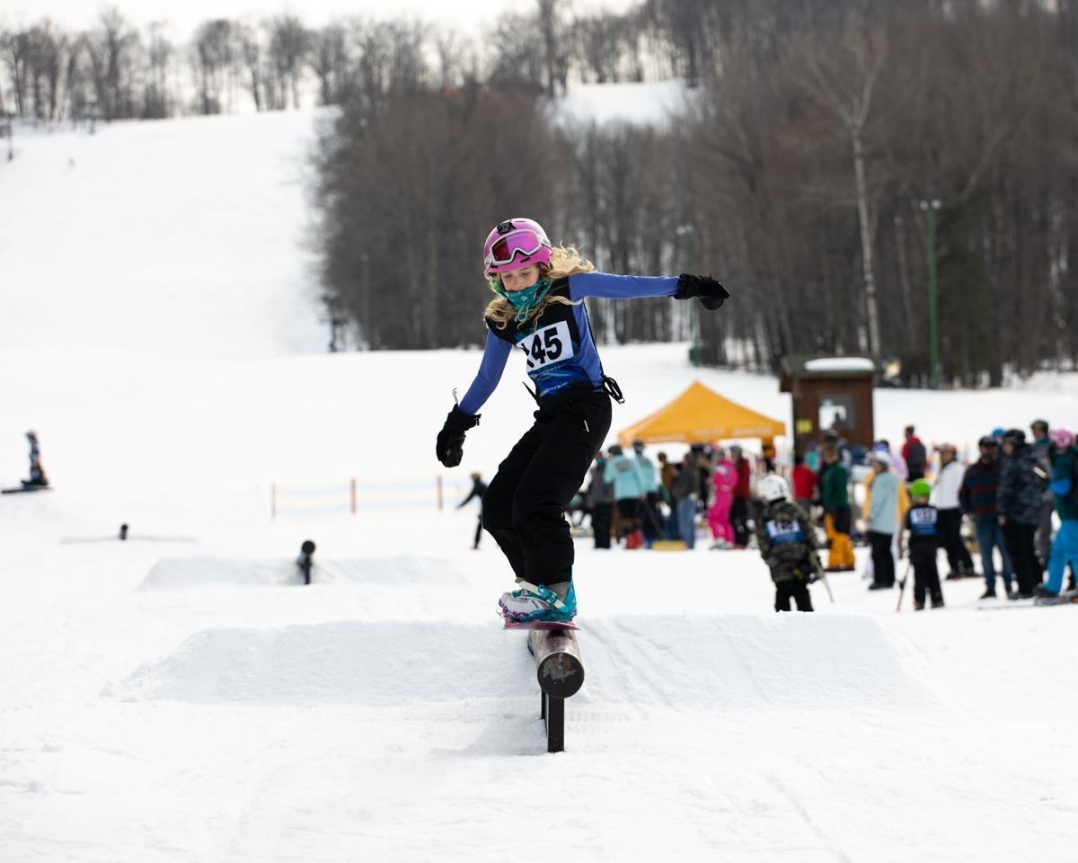 Snowboarder balancing on a rail with a snowy background, wearing a helmet and numbered vest.