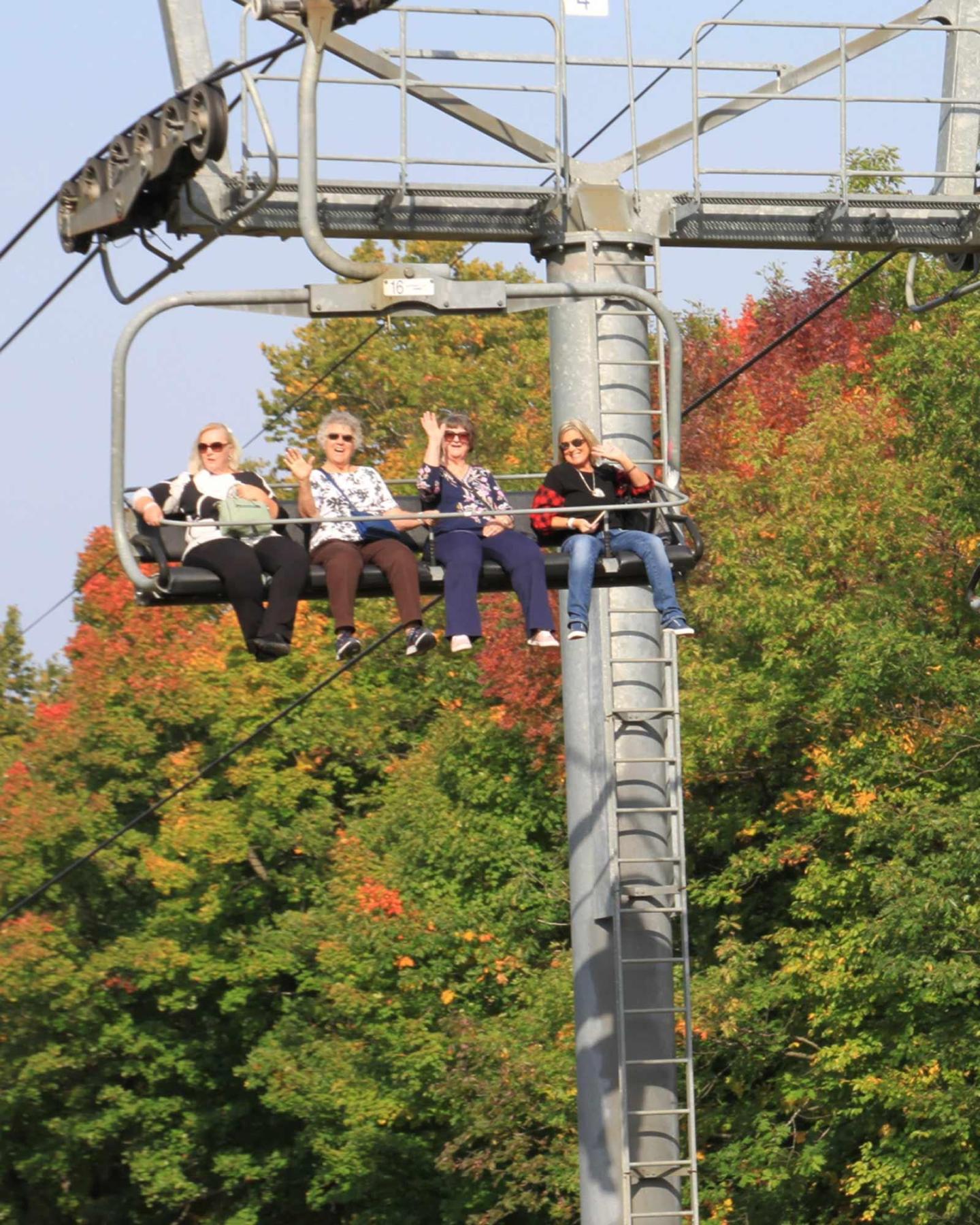 Four people on a ski lift against a backdrop of autumn trees.