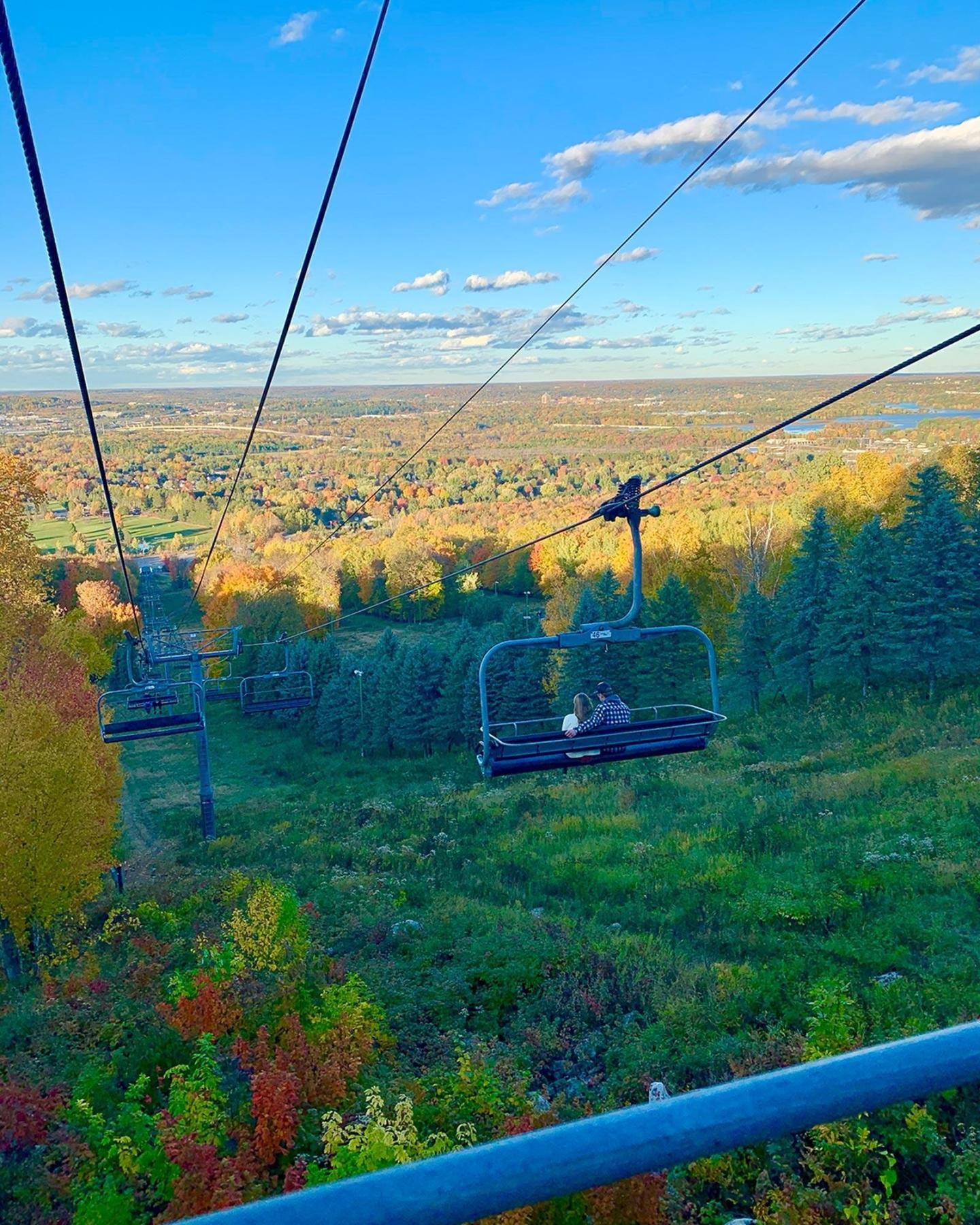Chairlift over a green hillside with colorful autumn trees and a clear blue sky.