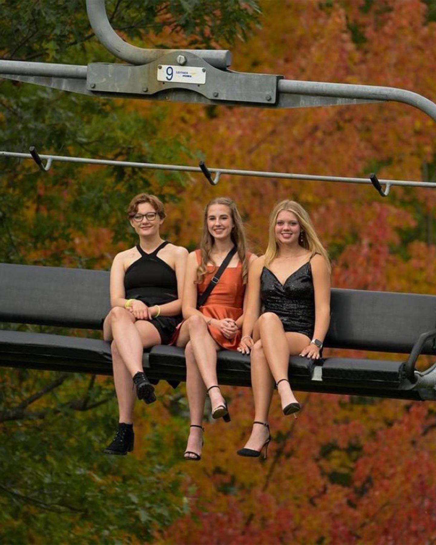 Three women in dresses on a chairlift with autumn leaves in the background.