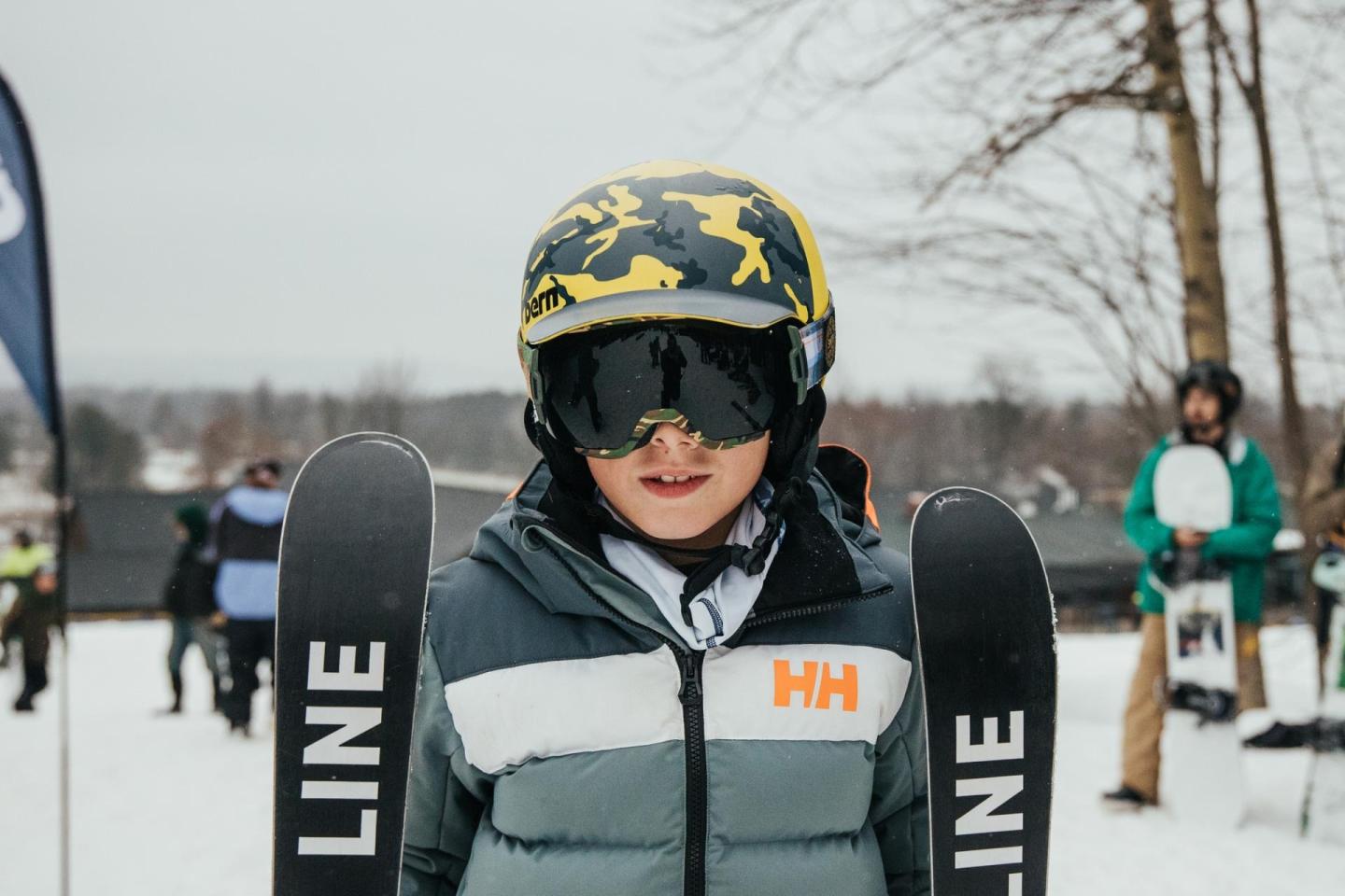Child skier wearing a helmet and goggles, holding skis in a snowy setting.