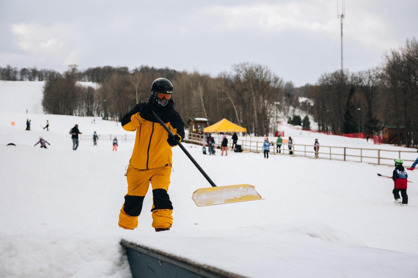Person in yellow snowsuit clearing snow on a ski slope, with others in the background.