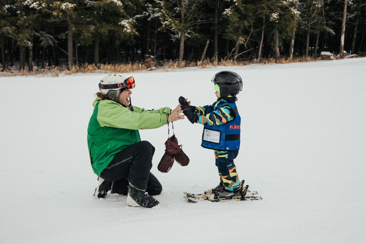 Adult kneeling on snowy field helping child in ski gear.