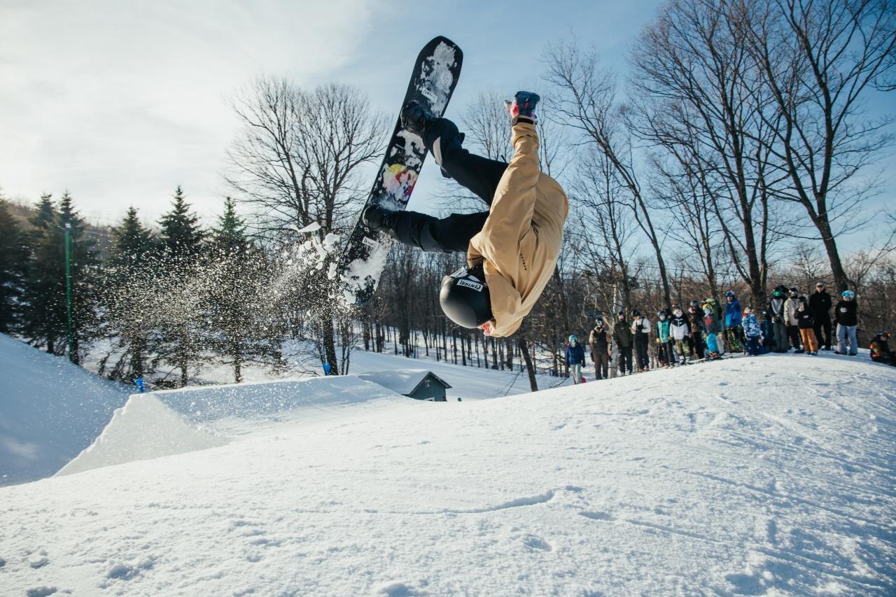 Snowboarder in mid-air doing a backflip on a snowy slope, surrounded by trees and spectators.