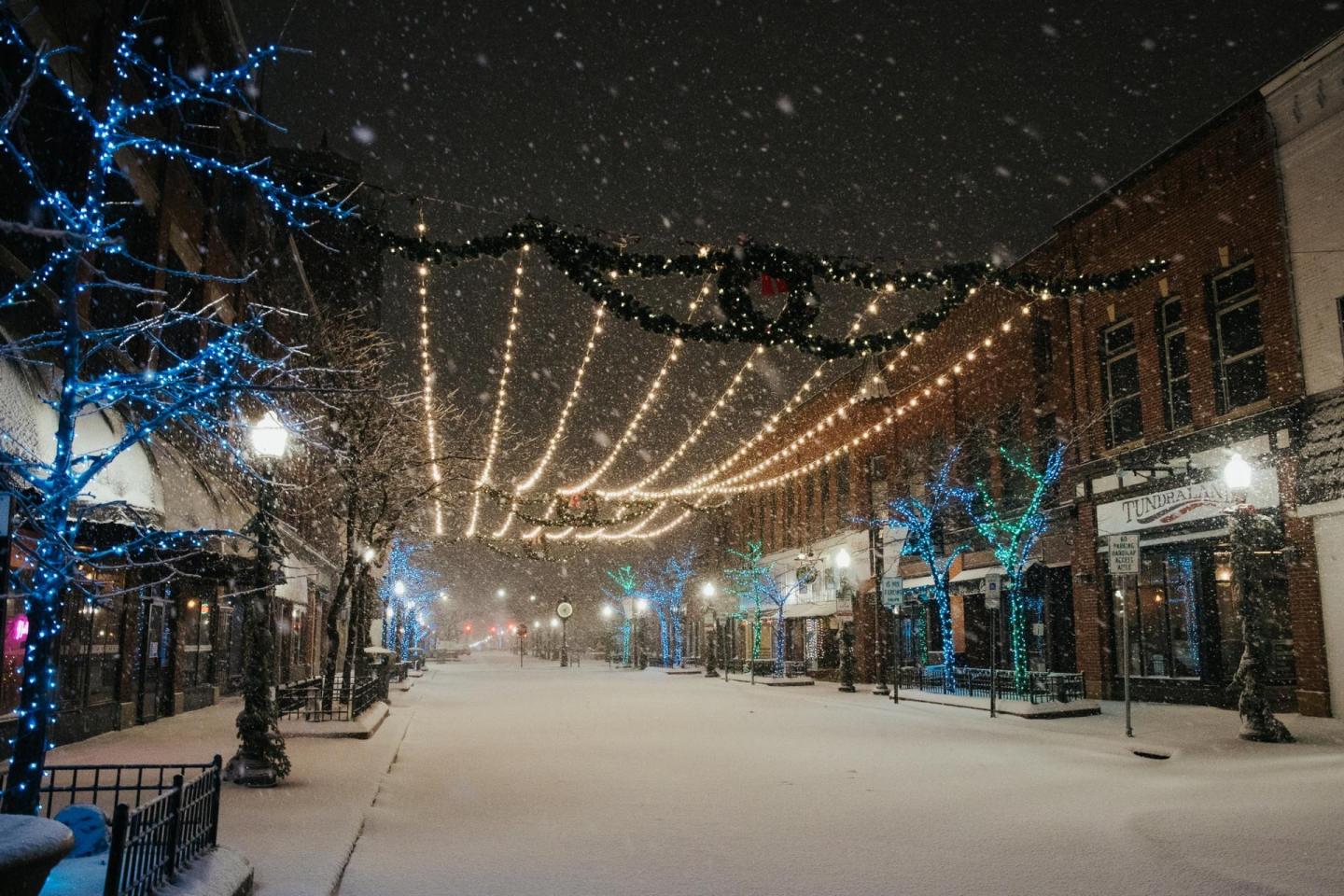 Snowy street at night with festive lights and blue-lit trees.