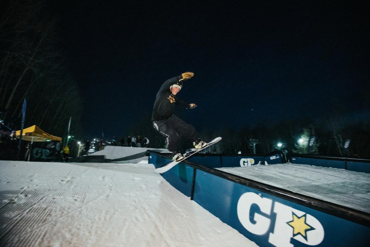 Snowboarder grinding a rail at night, surrounded by lights and trees.