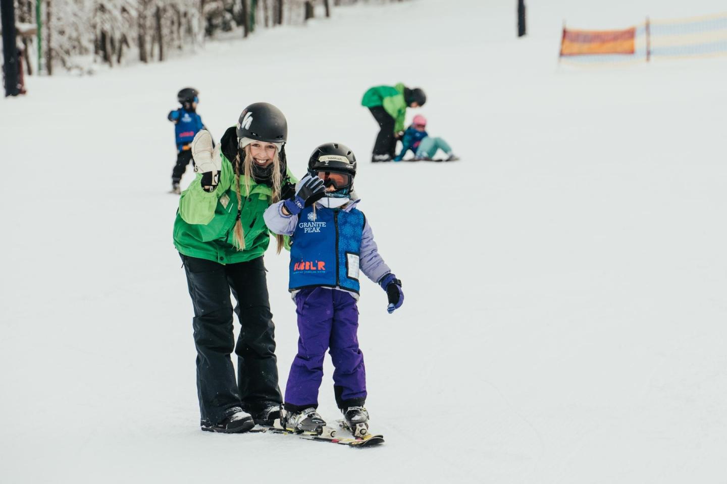 Woman and child skiing on snow, child waving. Others in background skiing.