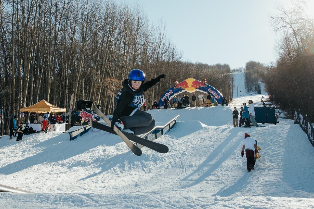 Snowboarder in mid-jump on snowy slope with trees and spectators in background.
