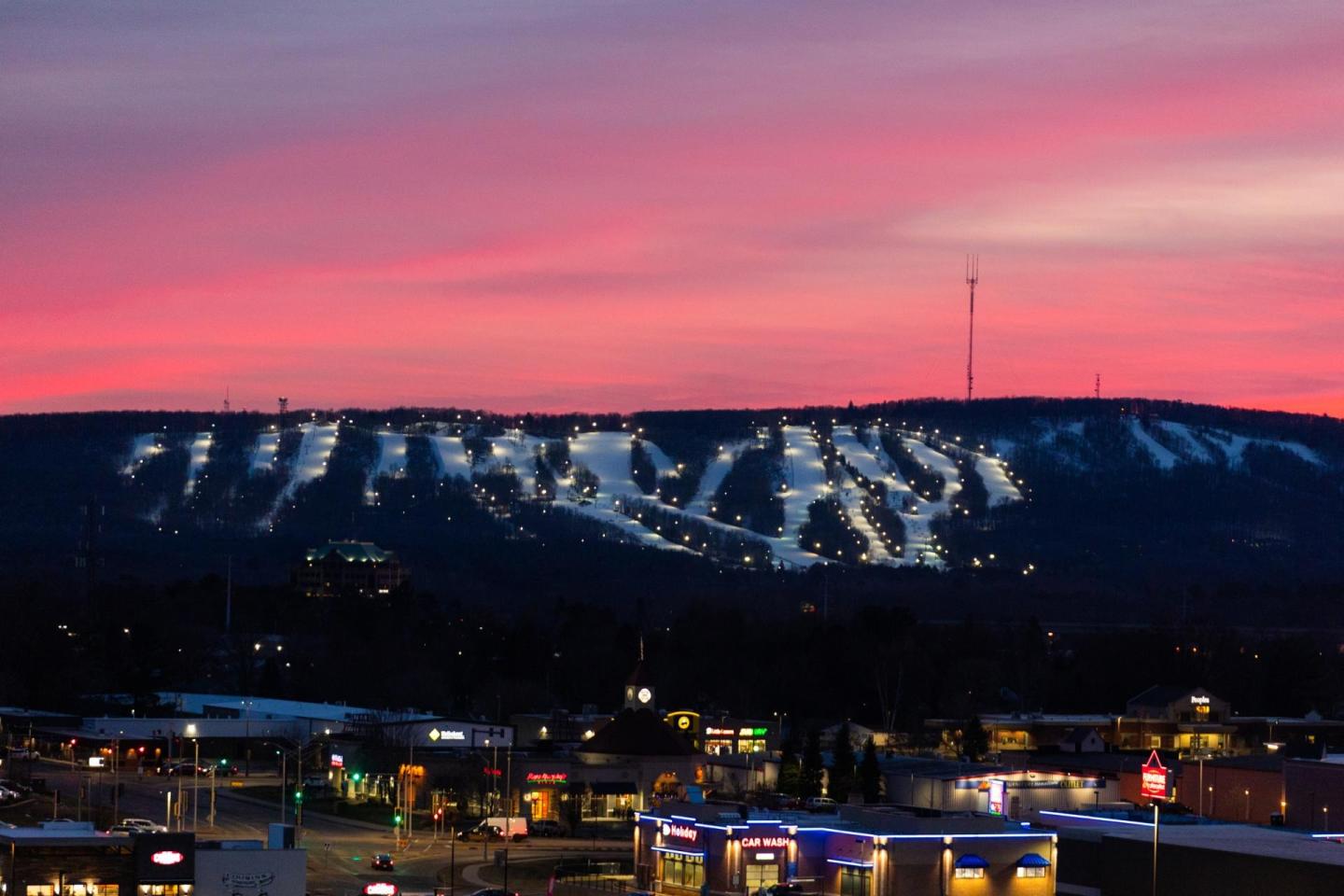 Cityscape with a pink sunset and illuminated ski slopes in the distance.