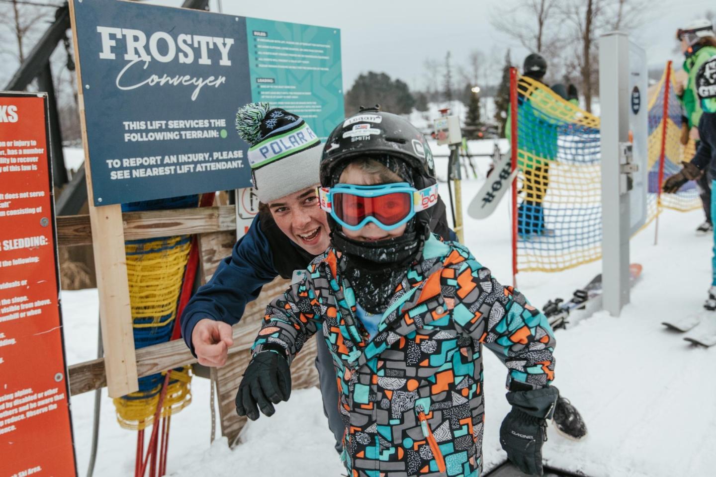 Young child in colorful snowsuit and helmet smiling on a snowy ski slope.