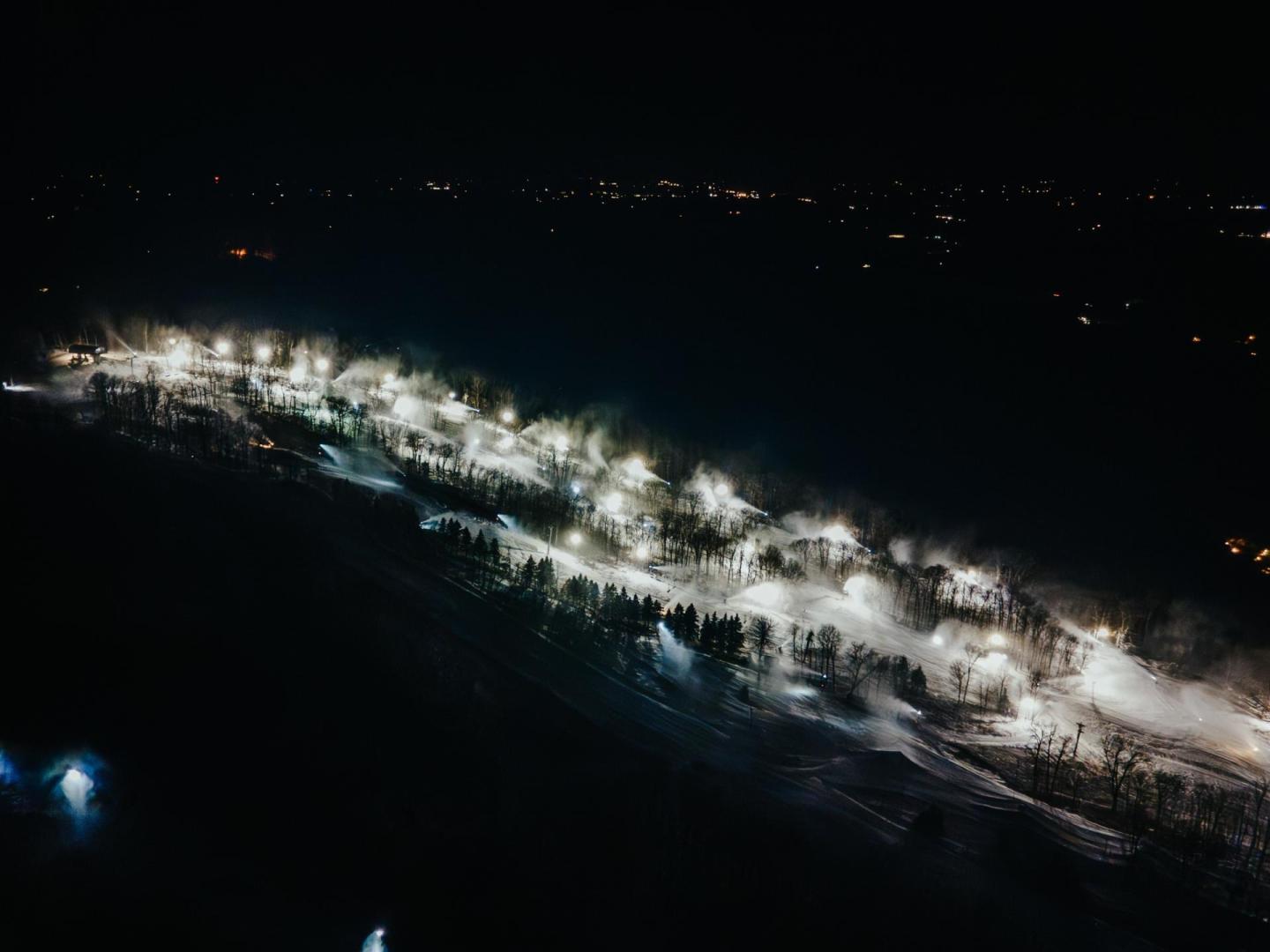 Nighttime ski slope illuminated, with trees and distant city lights.