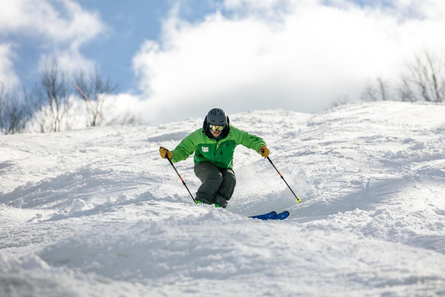 Skier in green jacket skiing downhill on snowy slope under blue sky.