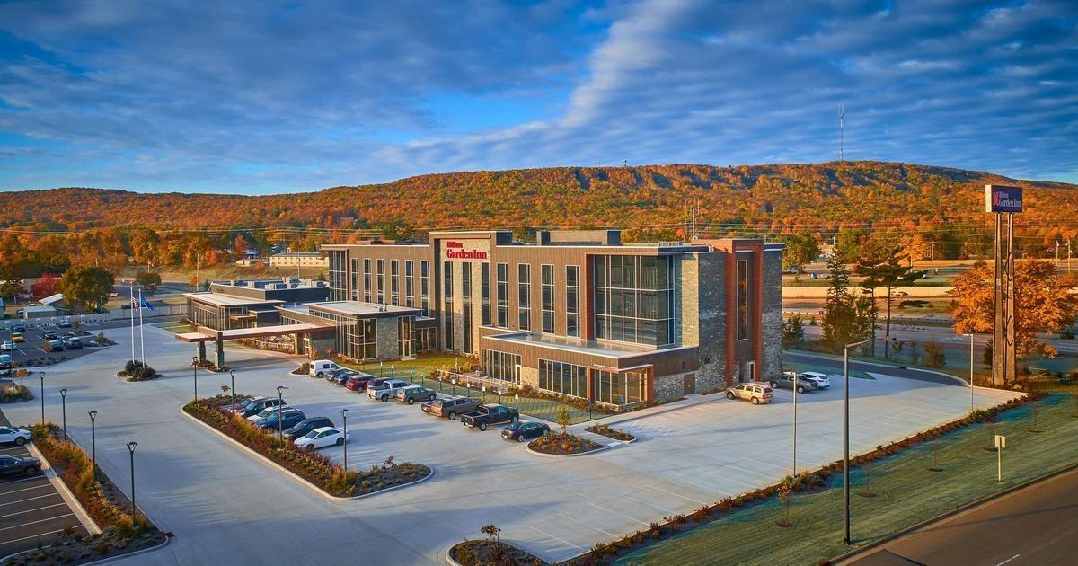 Hotel with parking lot, surrounded by autumn trees and hills under a blue sky.