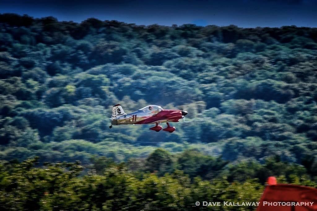 Red airplane flying over green forested hills.