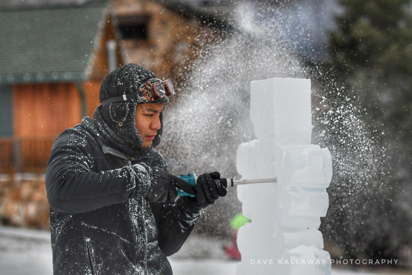 Man carving an ice sculpture, snow flying, wearing winter clothing.