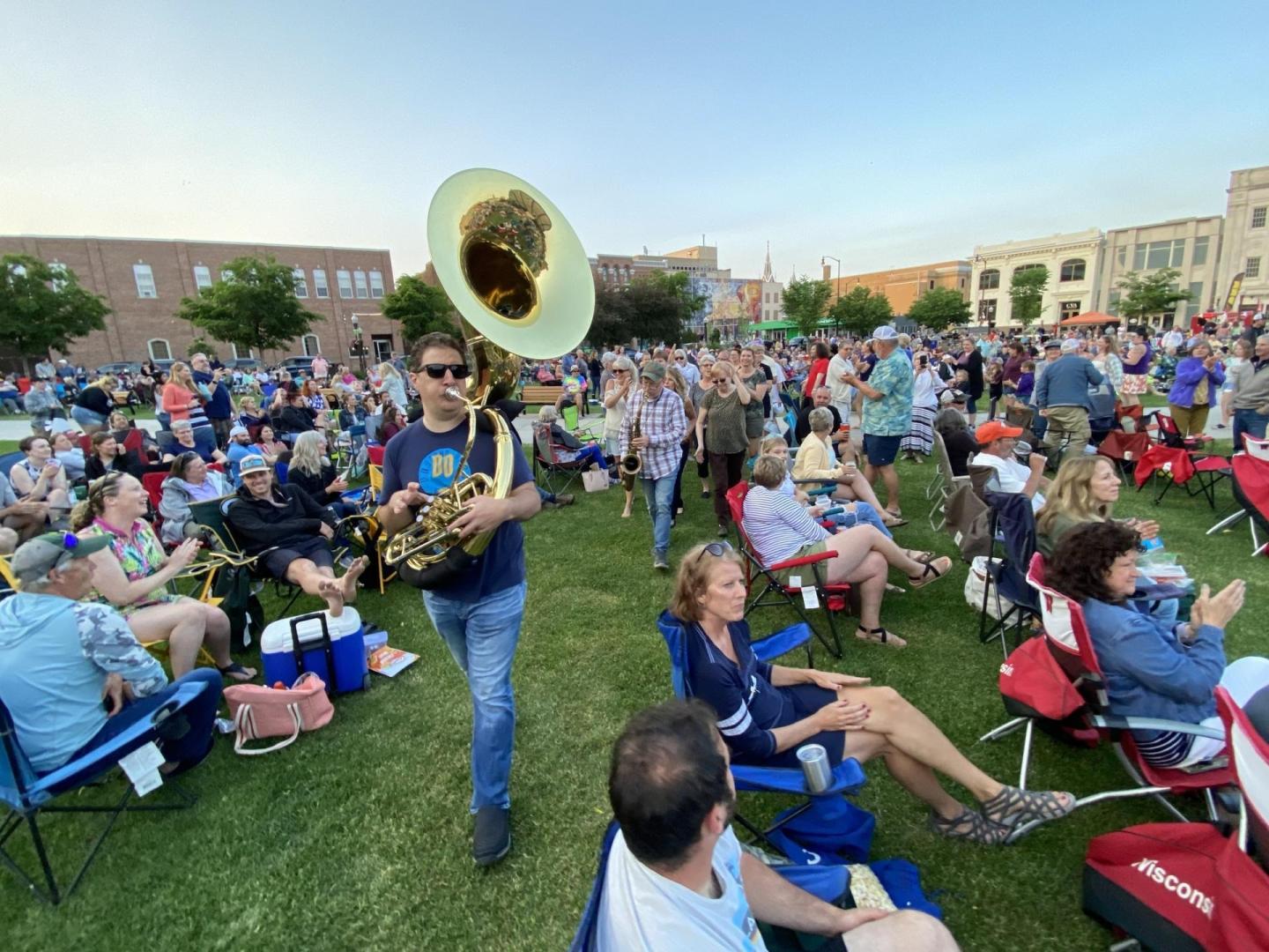 Outdoor crowd with tuba player walking through a seated audience on grass.